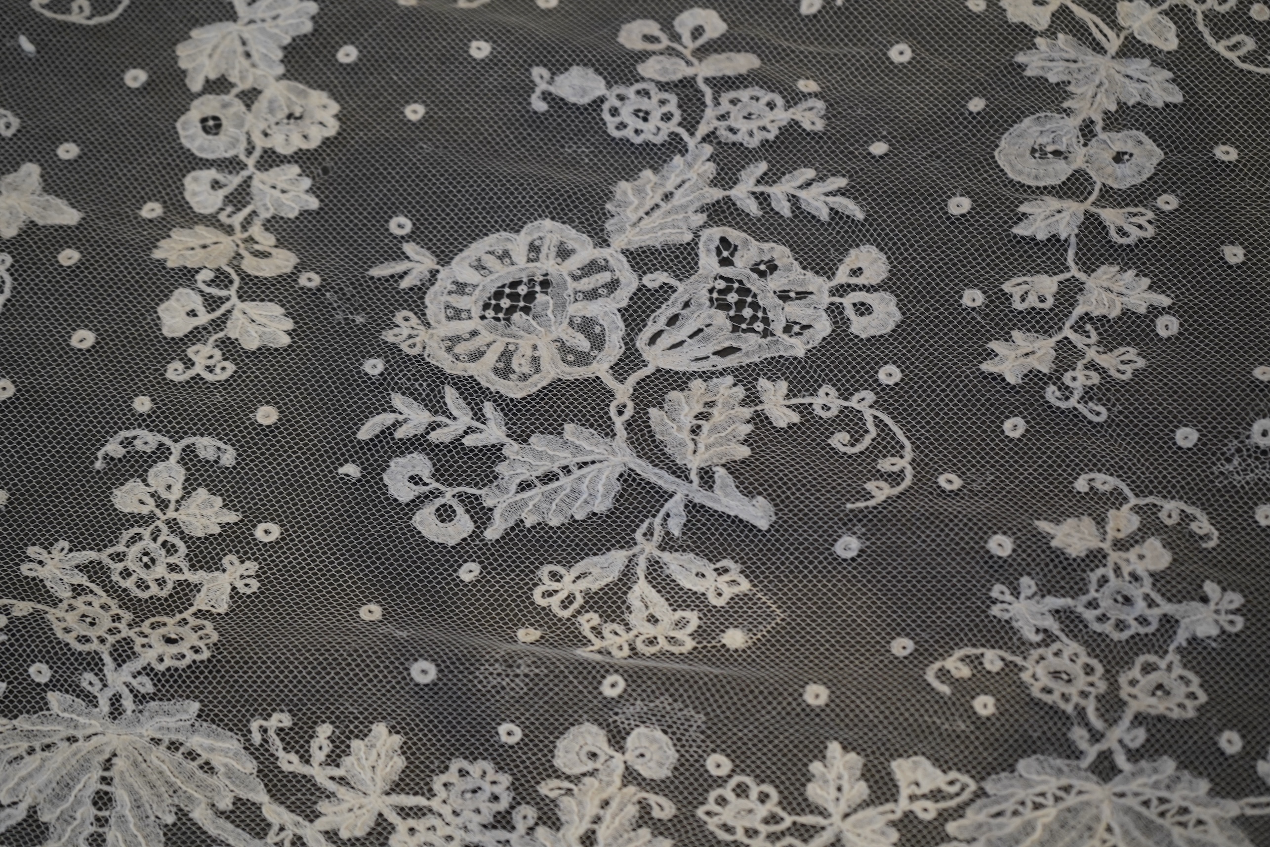 A deep elaborately designed flounce of mid 19th century Brussels bobbin appliquéd lace on net, with finished lace ends, worked with an intricate floral border below large floral cartouches framed in similar motifs and al